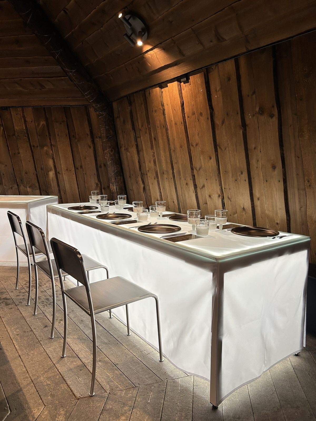 Megvilágított fehér asztal fém székekkel egy faházbelsőben az IWC privát rendezvényén, Longyearbyen, Spitzbergák