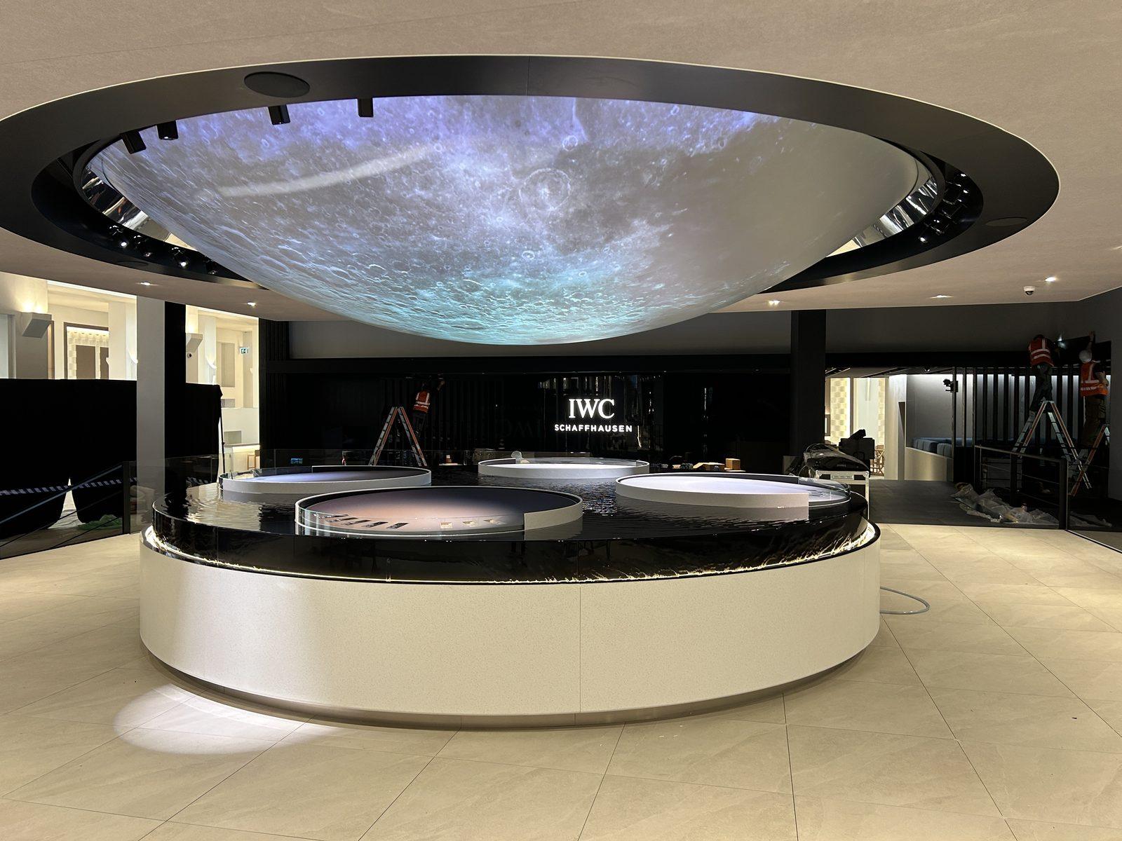 Kör alakú, vízzel töltött óra-display fehér talppal és holdfelszínt vetítő kupolával az IWC standján a genfi Watches & Wonders kiállításon